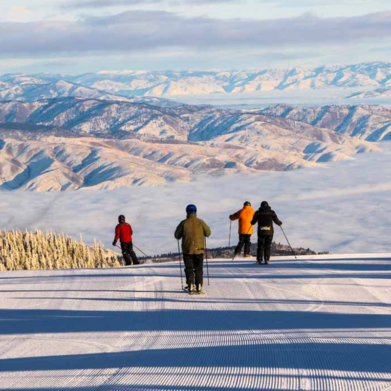 Group of skiers on fresh corduroy with cloud covered valley view below.