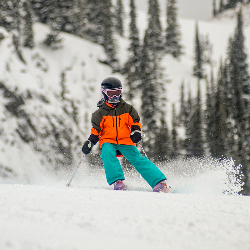 Young skier in oragne and black jacket with green pants making turn and spraying snow