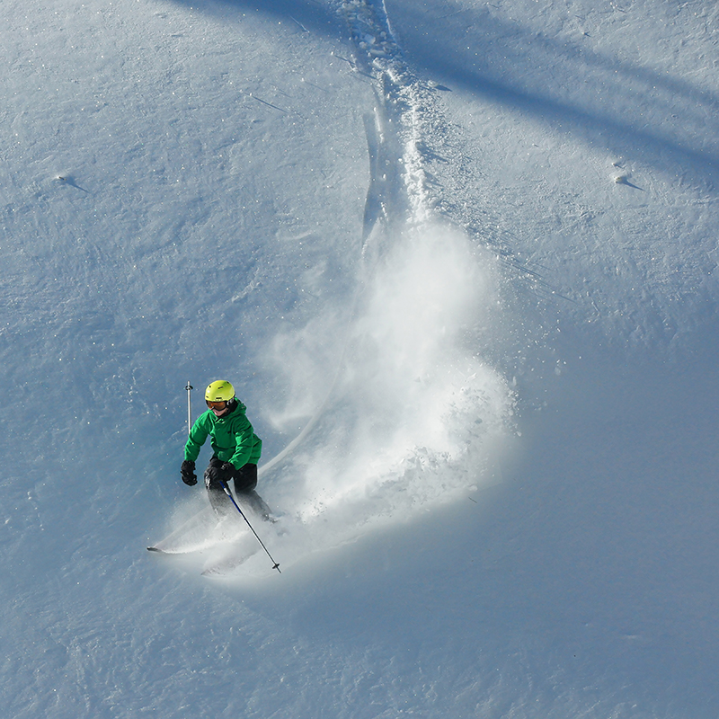 Skier in green jacket and yellow helmet making powder turn
