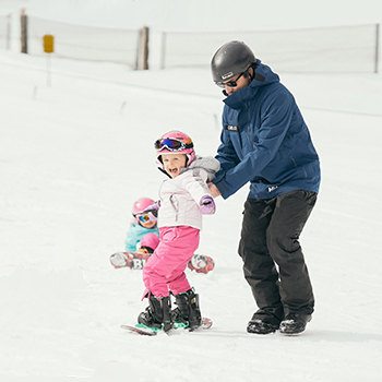 Instructor assisting a young snowboarding in pink pants, white jacket, and pink helmet