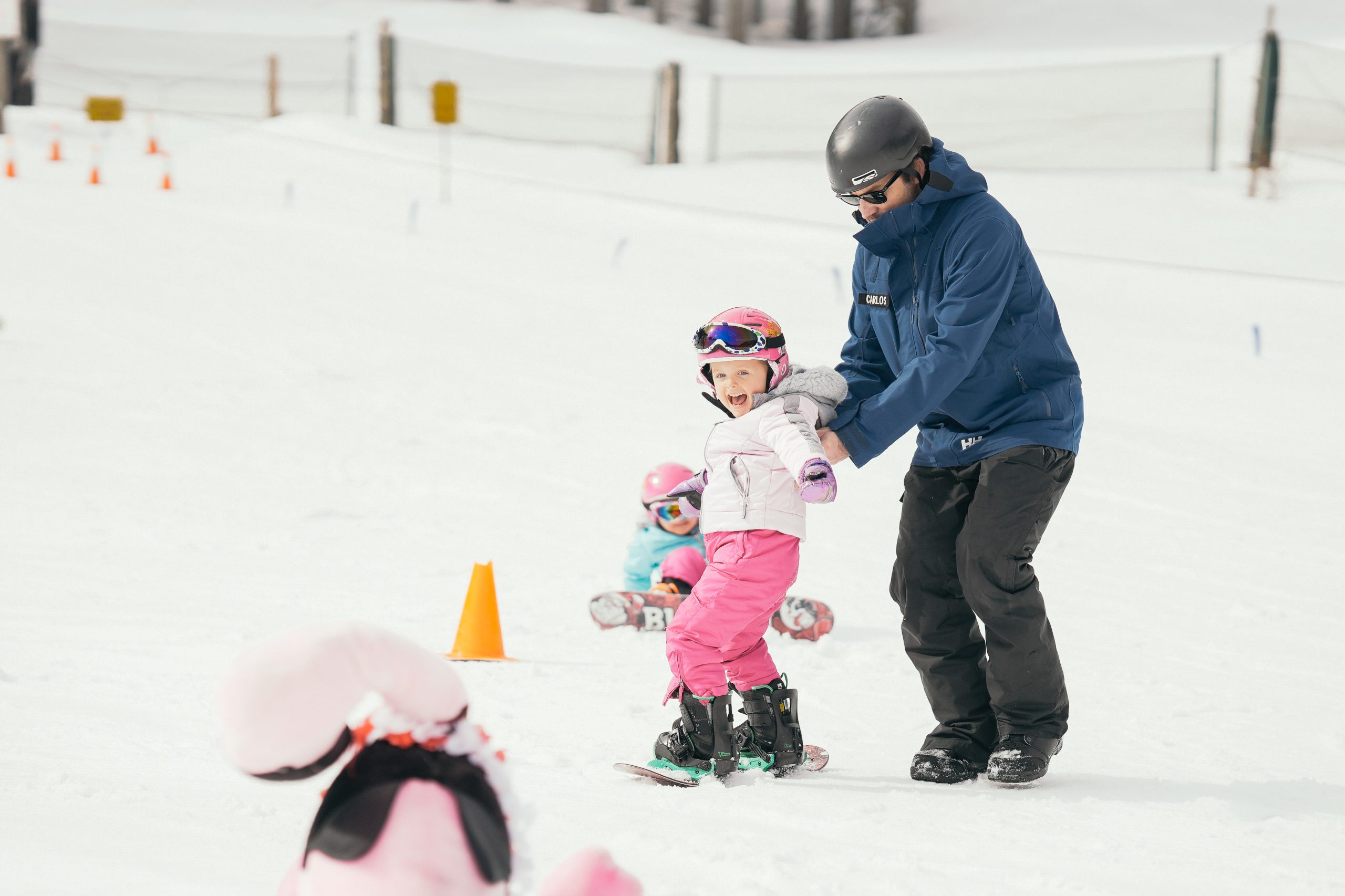 Instructor assisting a young snowboarding in pink pants, white jacket, and pink helmet