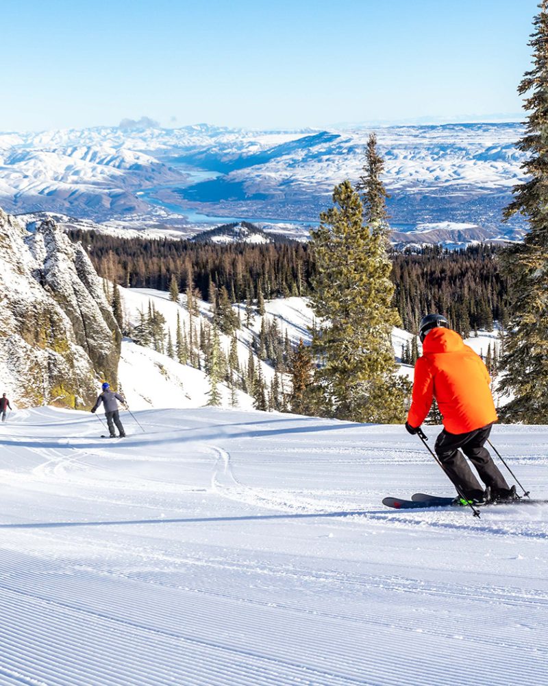 Skier in orange coat making turn on fresh corduroy with valley view below