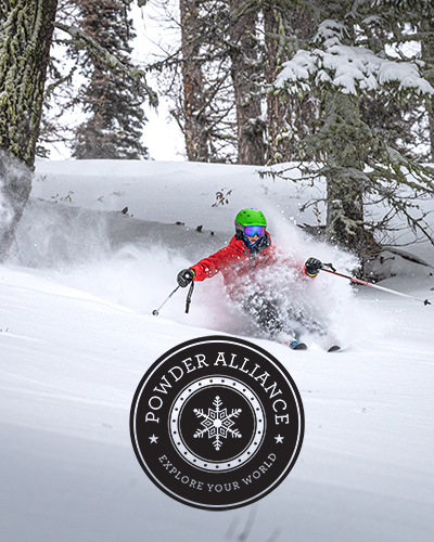 Skier in red jacket and green helmet making a powder turn with Powder Alliance logo in front