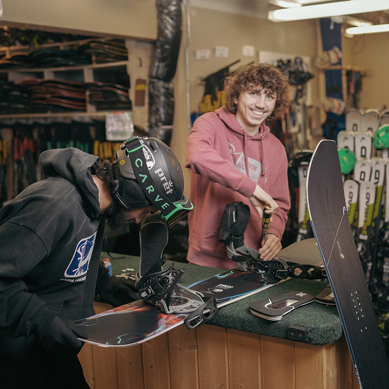 Rental technician in red smiling while adjusting a snowboard binding for a guest in black