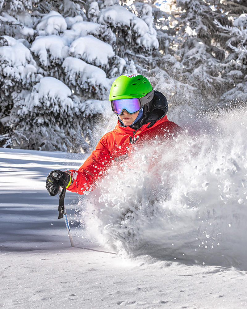 Young skier in red jacket and green helmet deep in a powder turn