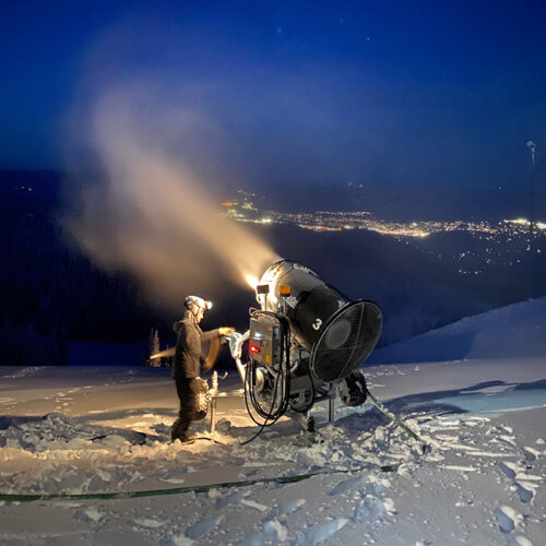 Snowmaker working with a snow gun at night with city lights below