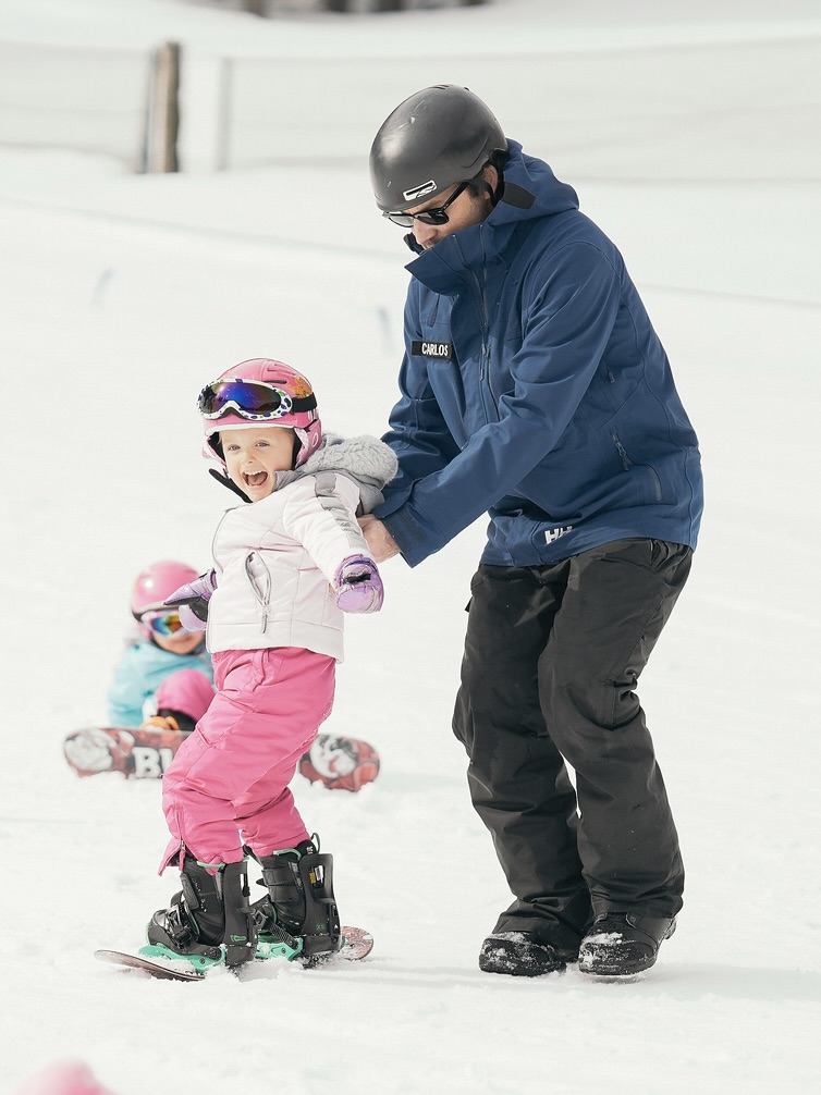 Instructor assisting a young snowboarding in pink pants, white jacket, and pink helmet