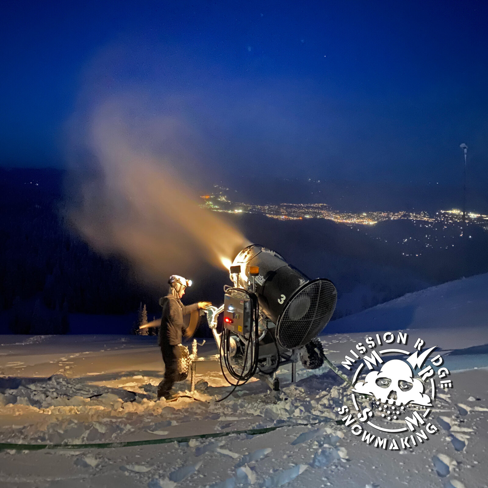 Snowmaker working with a snow gun at night with city lights below