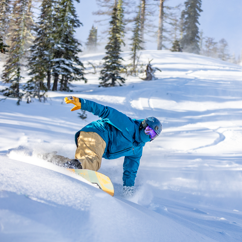 Snowboarder in blue jacket making hard carve with tree and cliffs behind.