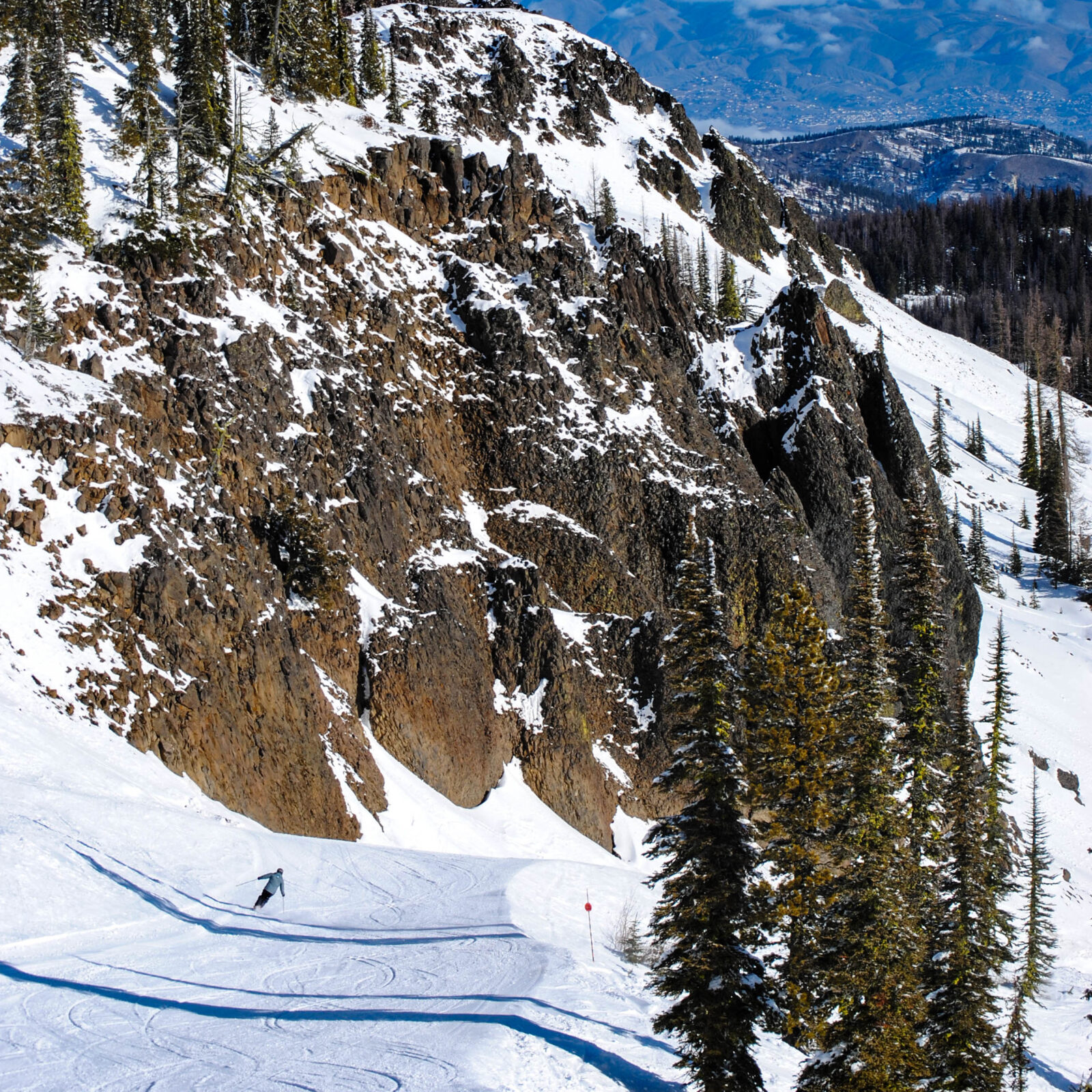 Skier on a sunny day skiing down Bomber trail at Mission Ridge with the Bomber Cliffs and the Wenatchee Valley in the background.