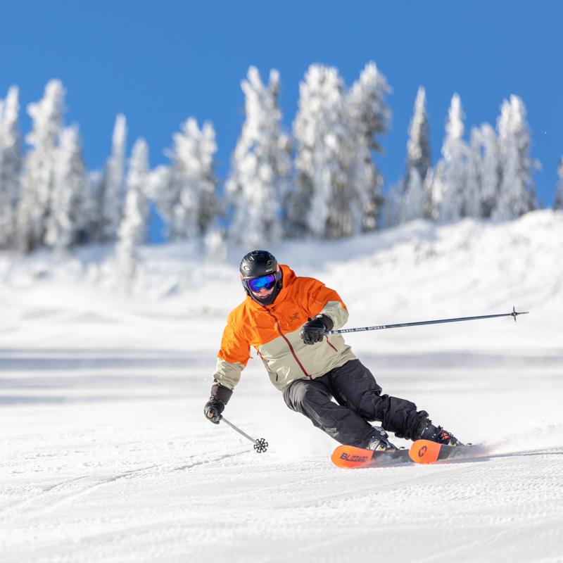 Skier in an orange and white jacket and black pants making a turn on a groomed run at Mission Ridge with blue sky and snowy trees in the background.