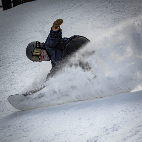 Snowboarder in black throwing snow as he carves under chair 3
