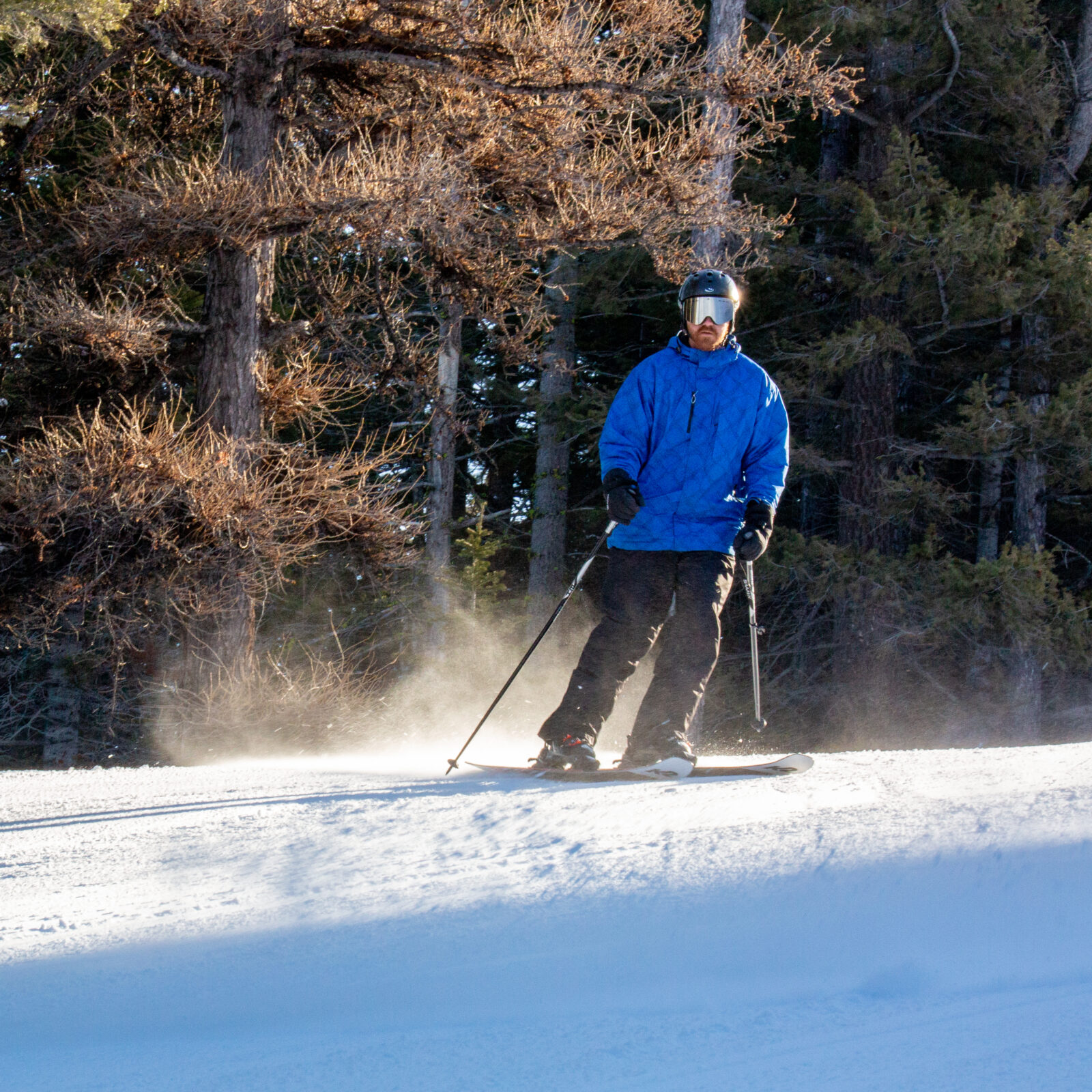 Skier in blue jacket goes down Sitkum run on Chair 4 at Mission Ridge. Some snow sprays behind the skier in the sunlight.