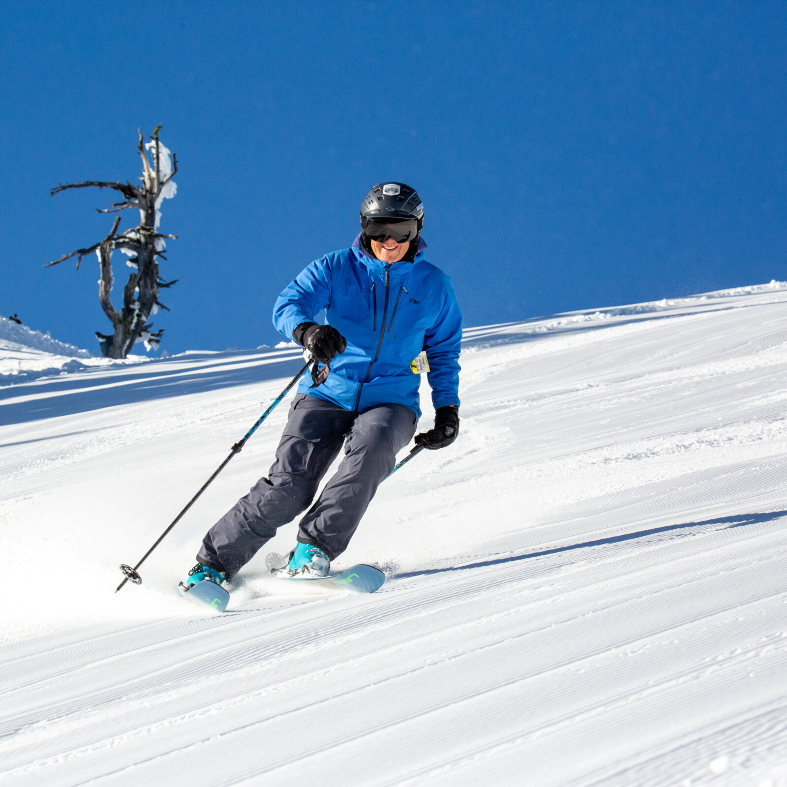 Skier in blue jacket and grey pants goes down Katsuk with interesting tree in background. The sun is shining and the run has untouched corduroy. Chair 2 at Mission Ridge.