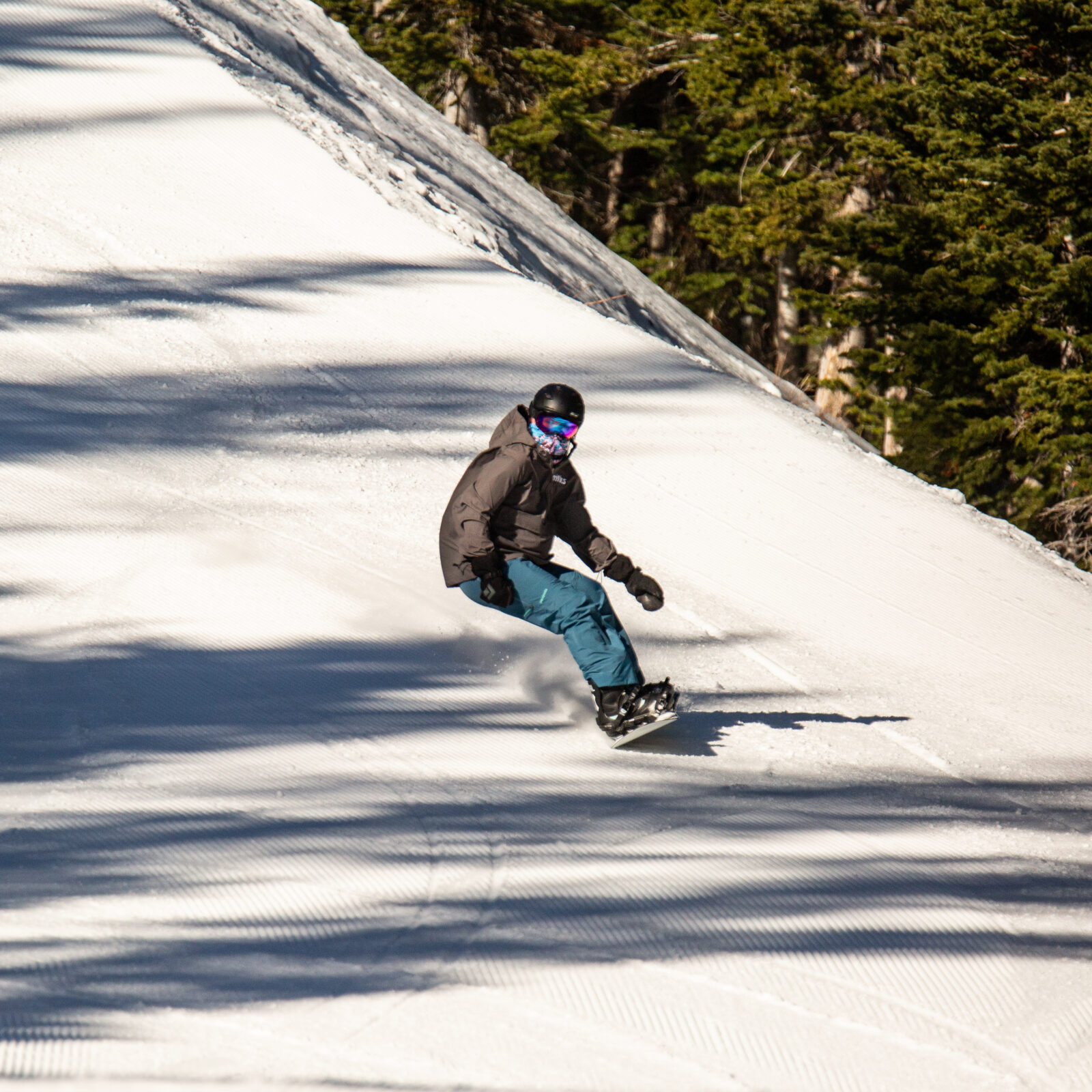 Snowboarder on Kiwa trail off of Chair 3 at Mission Ridge. He is wearing blue pants and a grey jacket. It is a sunny day with some shadows from trees.