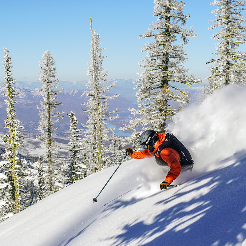 Skier in orange jacket and black vest making deep powder turn at Mission RIdge with river view below.