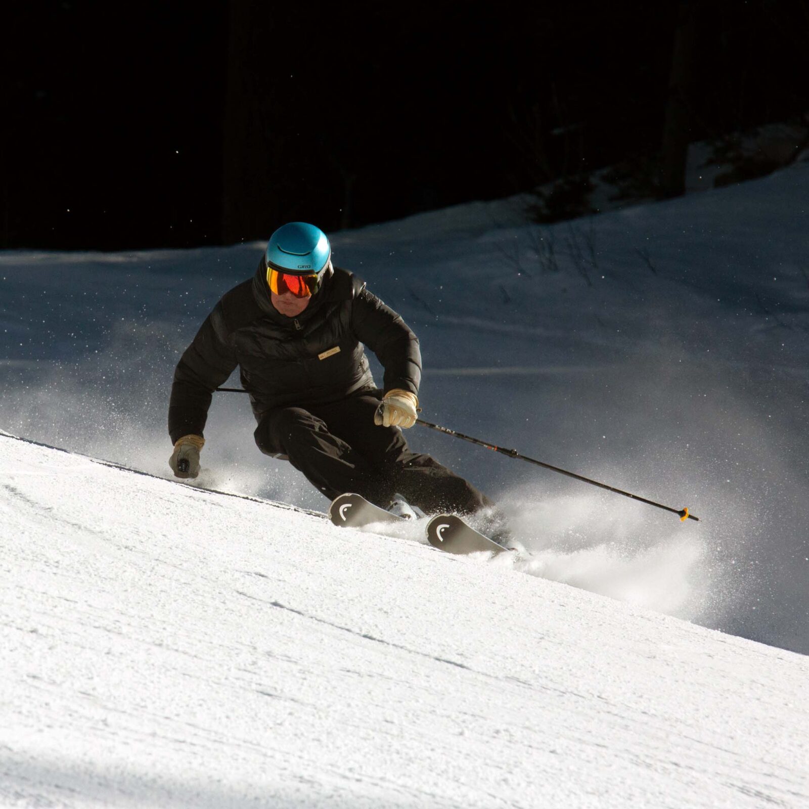 Skier in black jacket and blue helmet carving an aggressive turn and spraying snow.