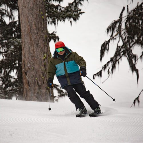 Mission Ridge employee Tom skis down Kiwa wearing a red helment and multi colored jacket. There are some mossy trees in the background. This run is off of Chair 3.