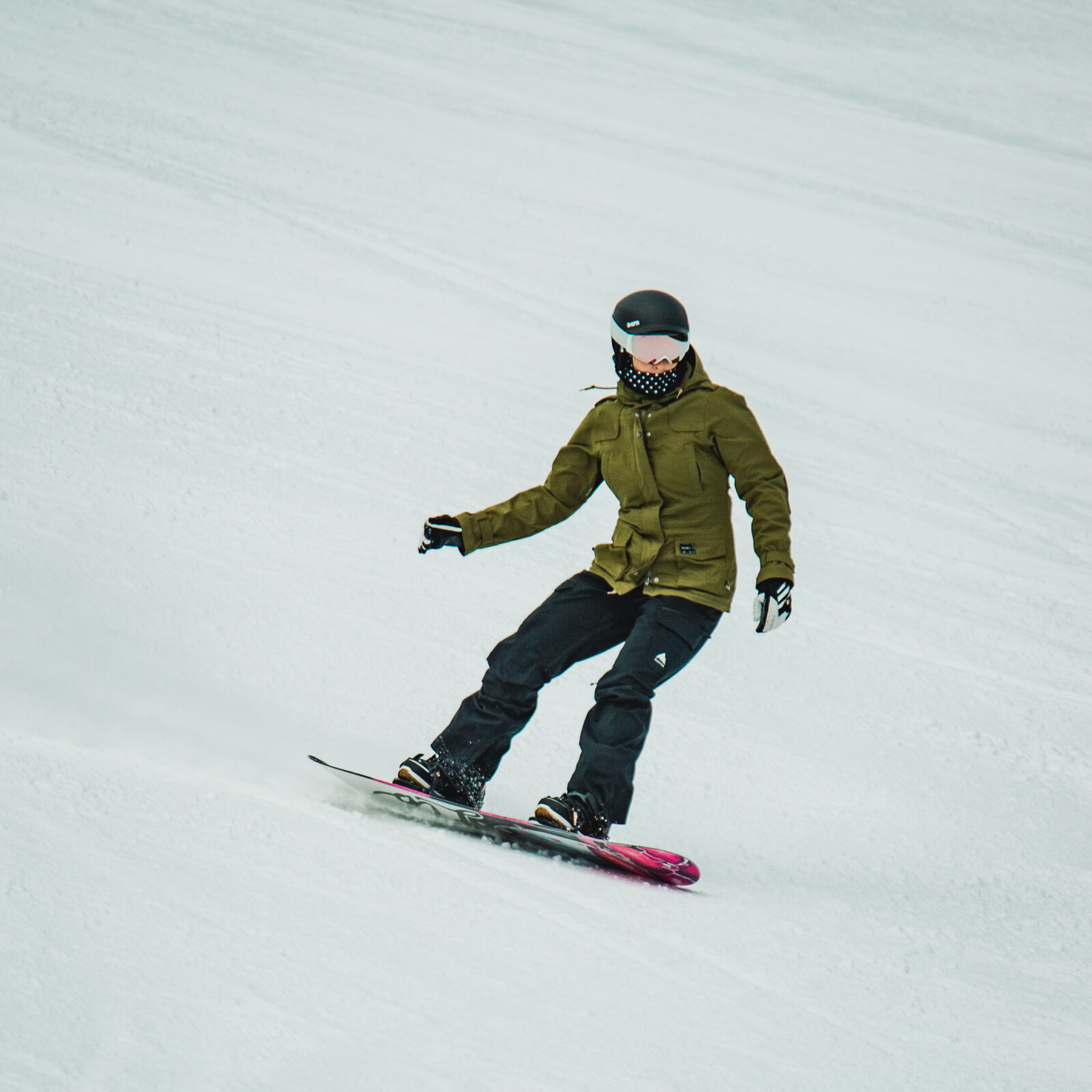 Women in green jacket and black pants snowboards down Tumwater 3 at Mission Ridge. This run is off Chair 2. Some snow sprays behind her.