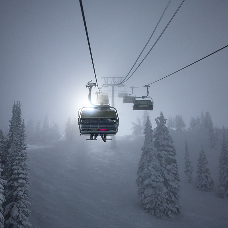 A dramatic winter image featuring a ski chairlift ascending into a misty, snow-covered landscape. The chairs are illuminated by a bright light, creating a striking contrast against the subdued gray sky. Snow-laden evergreen trees line the slope below, adding to the serene and atmospheric mood of the scene.