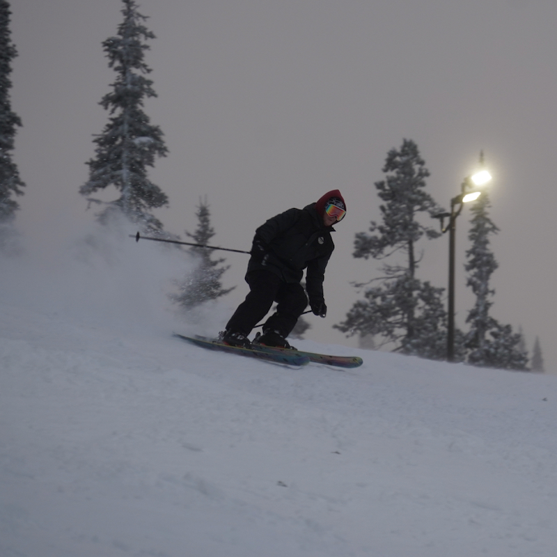 A skier descends the Tumwater ski trail at Mission Ridge Ski Area in the evening with a light on in the background.