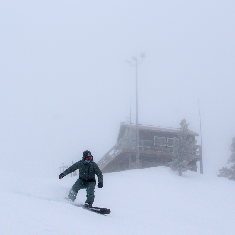 a snowboarder sends a powder spray on a gloomy day. a patrol shack is in the background.