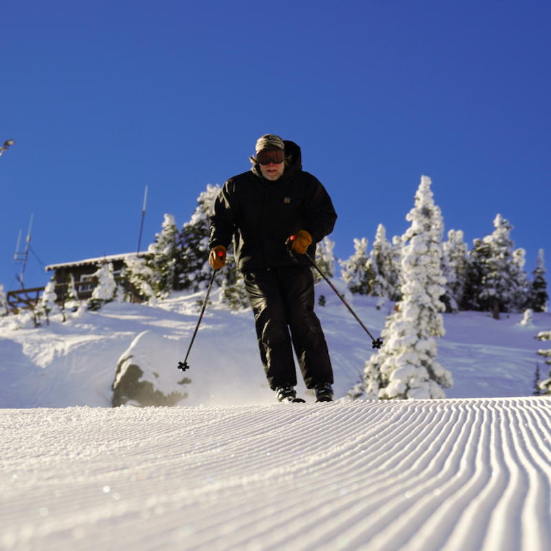 A skier descends the Sunspot trail at Mission Ridge on a bluebird day, with corduroy in the foreground and the ski patrol shack in the background.