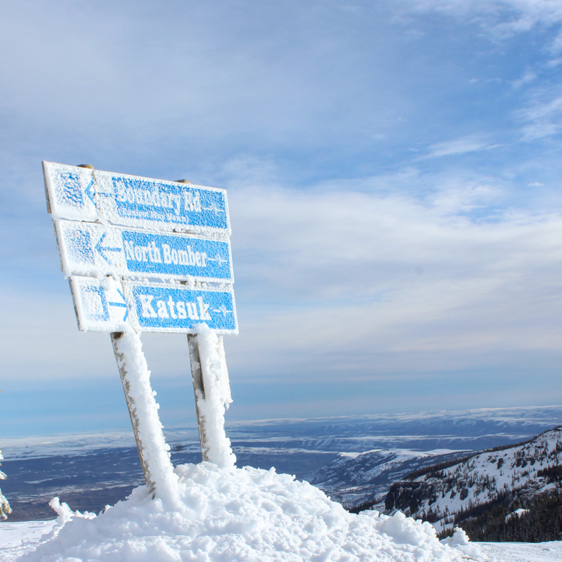 a rimey sign points to runs. it sits in a pile of chunky snow overlooking a sunny valley