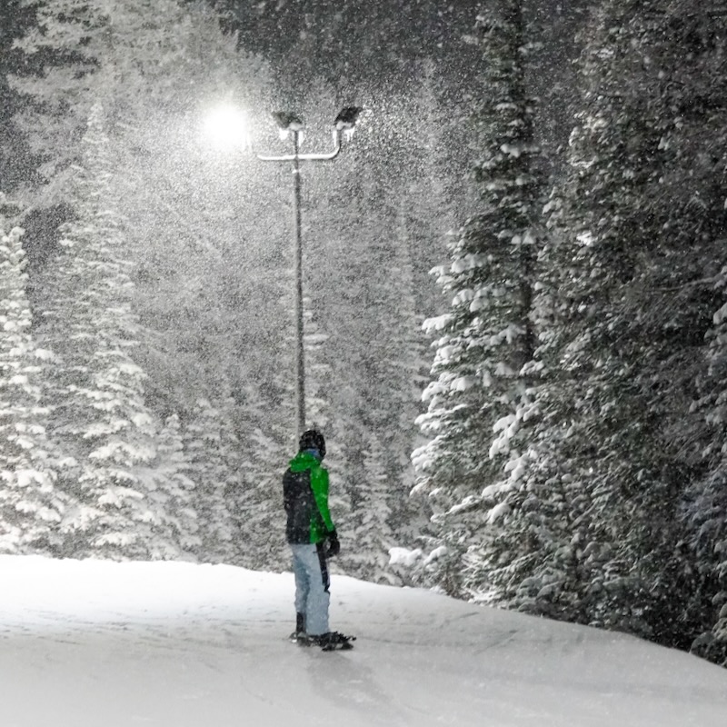 A skier traverses on a cat track at Mission Ridge in the night. There is a light behind him and it is snowing, creating a light background.