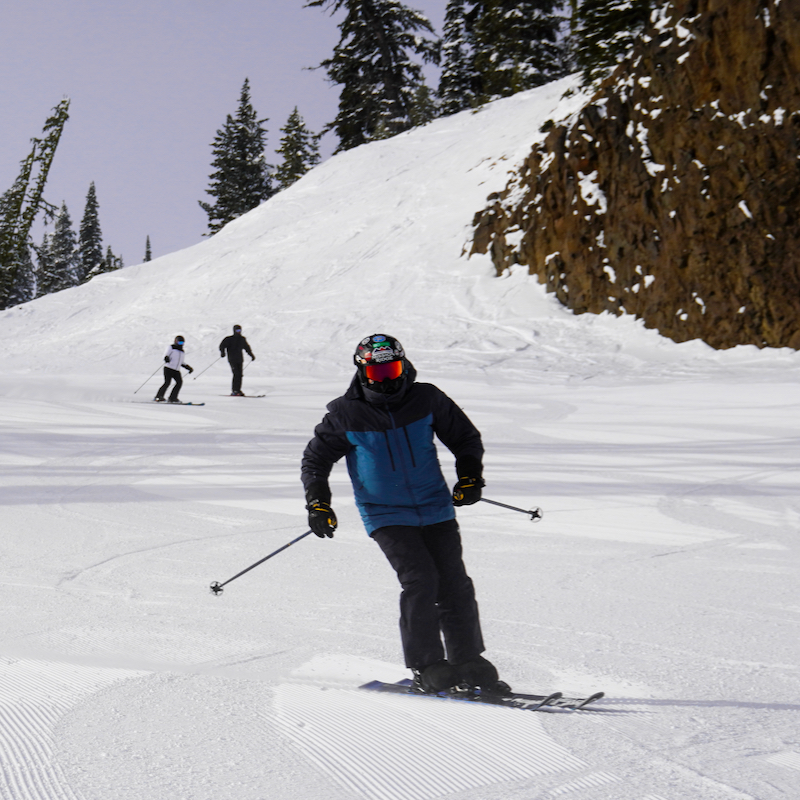 A skier descends the Bomber trail at Mission ridge, with 2 other skiers and the Bomber cliffs in the background.