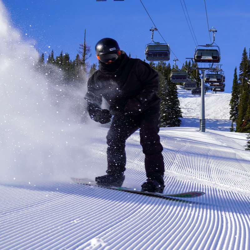 A snowboarder descends the Tumwater trail at Mission Ridge on a sunny day, spraying up a cloud of snow with the lift in the background.