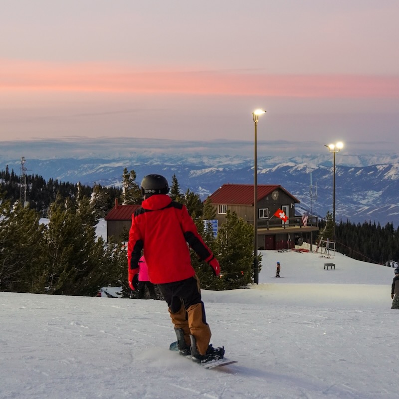 A snowboarder descends the sunspot trail at Moission Ridge, with a sunset in the background and the lights of night skiing.
