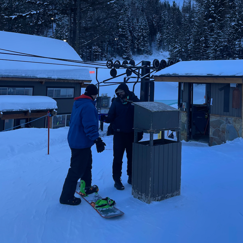 A ticket scanner scans a snowboarder's lift ticket at the bottom of Chair 1 at Mission Ridge in the late afternoon, with a building and the lift shack in the background.