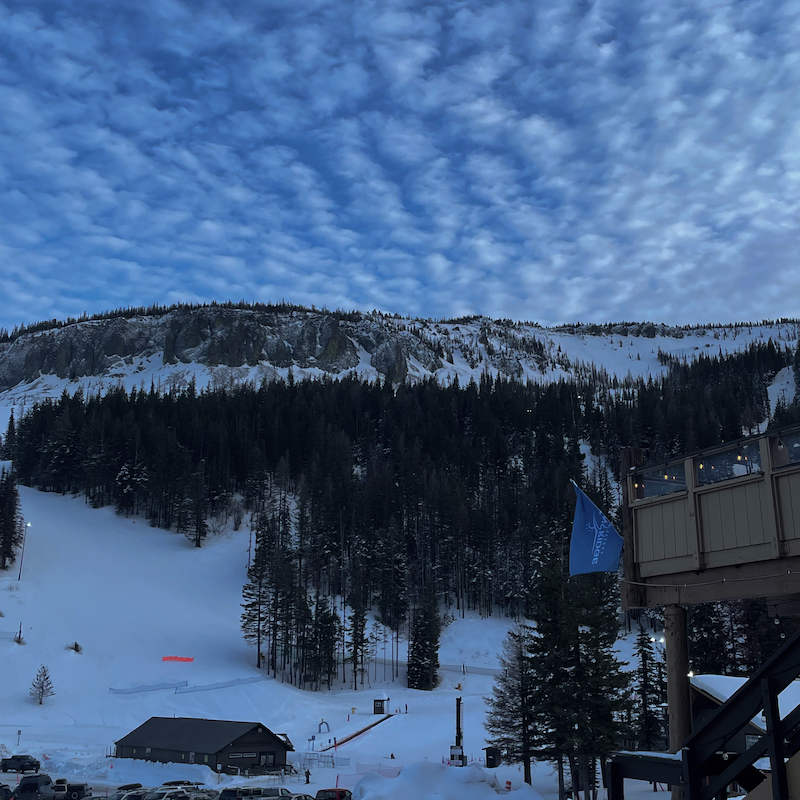 A view up the Lower Chak Chak run at Mission Ridge in the evening, with a corner of the lodge visible on the right, a sky checkered with clouds in the background, and a building, snowy ski run, and tree-covered hillside in the foreground.