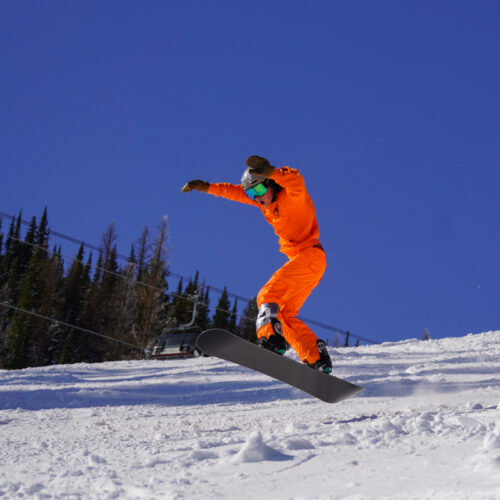 A young snowboarder descends the Nastar Trail at Mission Ridge, jumping in the air and raising his arms. He is wearing an orange outfit.