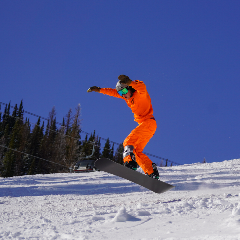 A young snowboarder descends the Nastar Trail at Mission Ridge, jumping in the air and raising his arms. He is wearing an orange outfit.