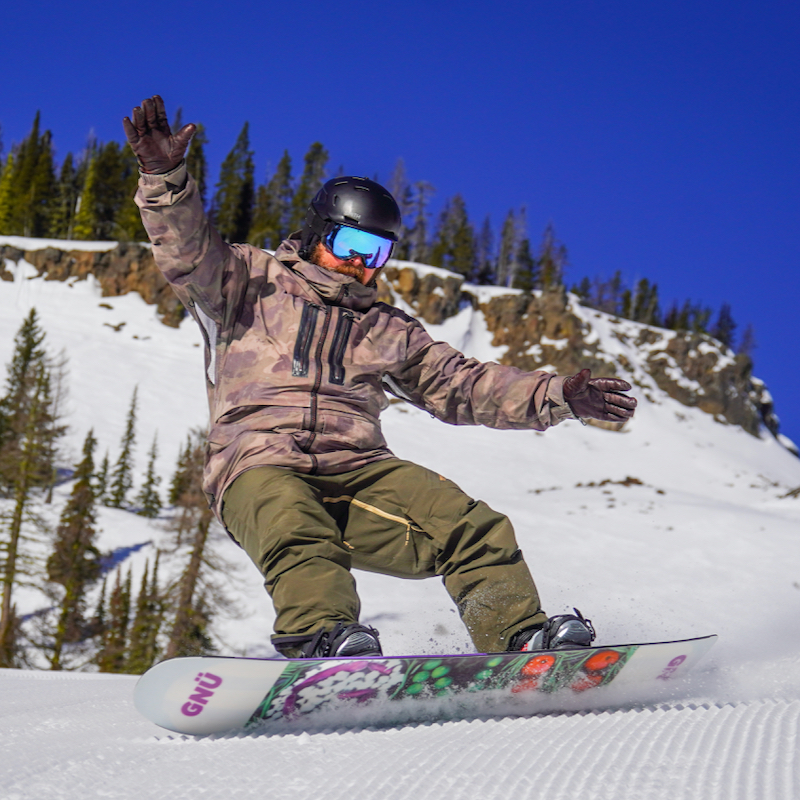 A snowboarder descends the Bomber Trail at Mission Ridge on a sunny day, with the Bomber Cliffs in the background.