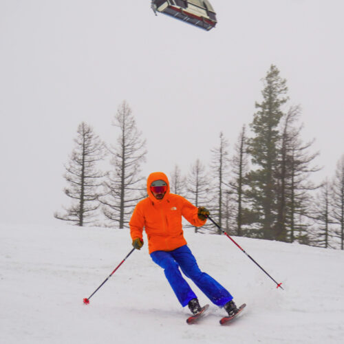 A skier descends the Tumwater Trail at Mission Ridge. He is wearing a bright orange jacket and blue pants.