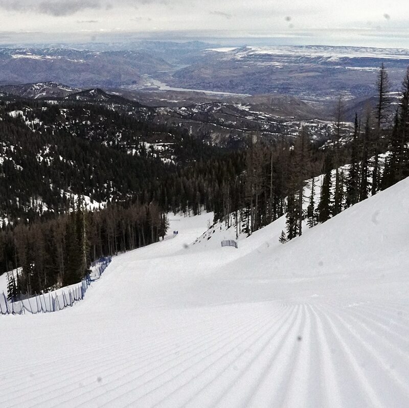 A stunning view of a freshly groomed ski run overlooking a vast mountainous landscape. The corduroy snow surface stretches down the slope, flanked by safety fencing and tall evergreen trees. In the distance, rolling hills and a winding river create a breathtaking contrast to the snowy foreground, showcasing the beauty of the alpine environment.