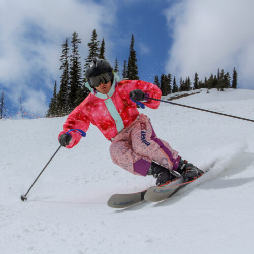 A skier in a pink onesie descends a trail at Mission Ridge, with snow, trees, and sky in the background.