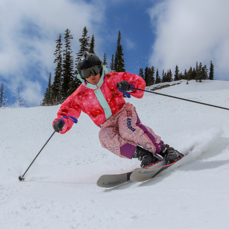 A skier in a pink onesie descends a trail at Mission Ridge, with snow, trees, and sky in the background.
