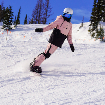 A snowboarder in a pink jacket descends a snowy slope with blue skies in the background.