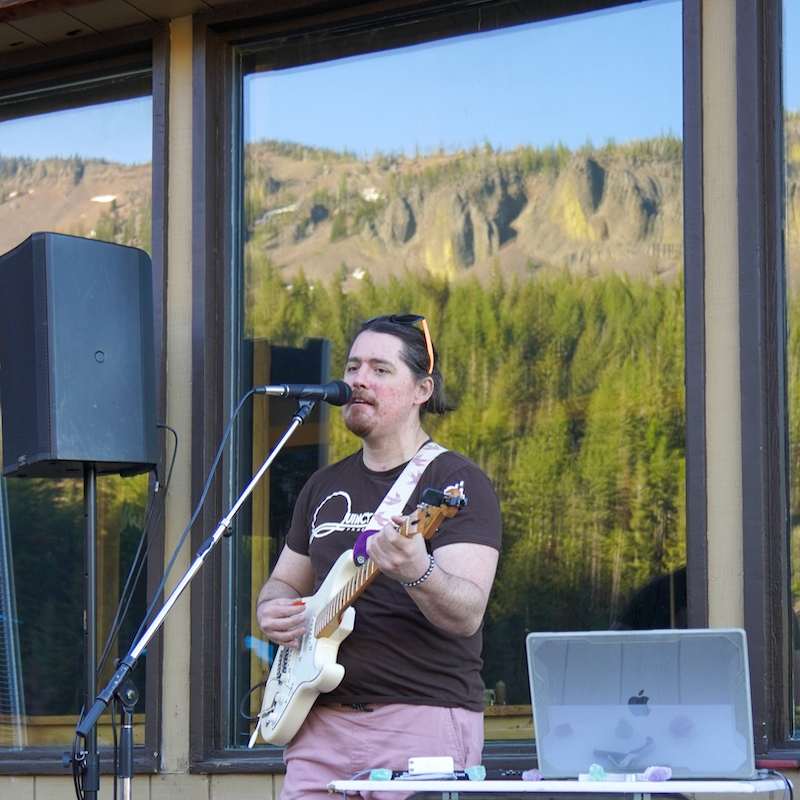 Local musician Micah J plays his guitar and sings into the microphone on the Hampton Lodge Deck, with a Mountain View reflected in the background.