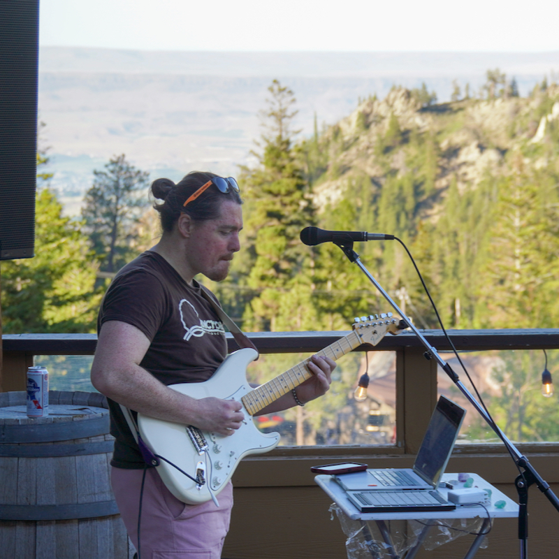 A singer plays an electric guitar on a wooden deck, with a view below looking down at a forested slope.