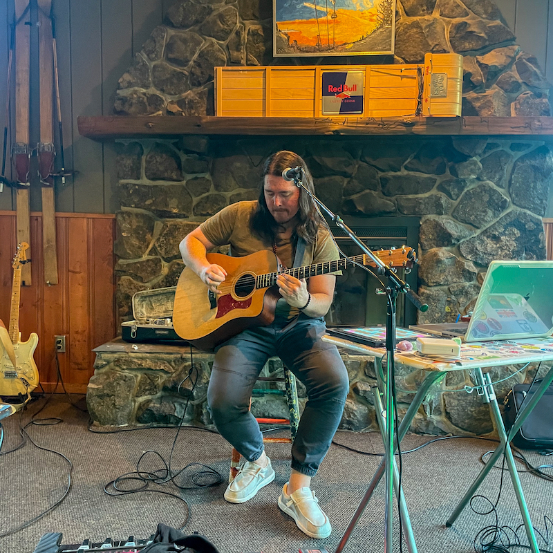 Local musician Micah J plays his guitar and sings into the microphone in front of the Hampton Lodge fireplace.