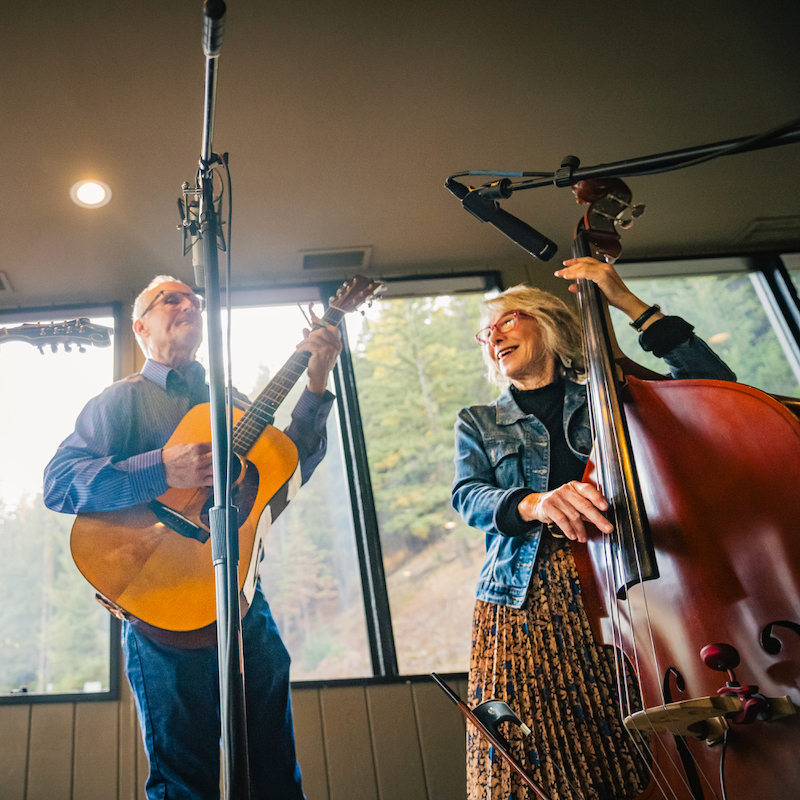 A man plays a guitar and a woman plays a bass in a room, with a window in the background showing a forest.