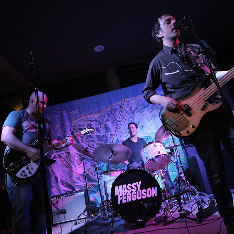 A high-energy live performance by the band Massy Ferguson, featuring three musicians on stage: a guitarist, a drummer, and a bassist/vocalist. The drummer’s kit prominently displays the band’s name on the bass drum. The backdrop features a Mission Ridge trail map, indicating the mountain venue. Colored stage lights and instruments create a vibrant, intimate atmosphere, perfect for a mountain-side music night.