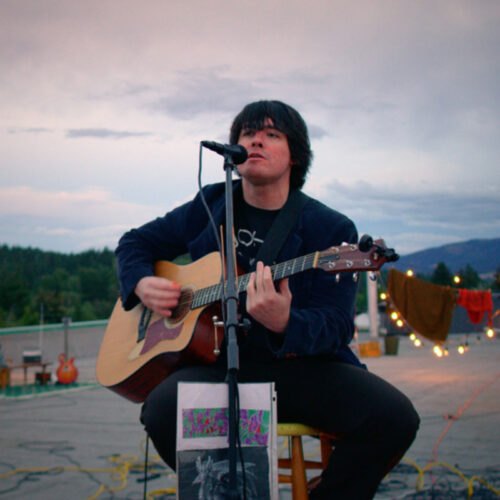 A musician performs at an outdoor venue during sunset, playing an acoustic guitar and singing into a microphone. The setting has a relaxed, creative vibe, with string lights, colorful decor, and mountains in the background. The sky is painted in soft hues of pink and purple, adding to the atmosphere of the intimate live music experience.