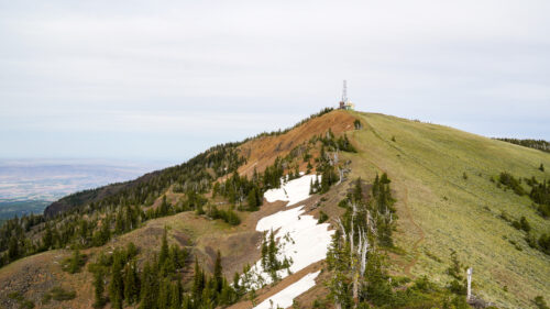a mountain vista showing a slope composed of a mixture of rocks, grass, and trees, with a couple snow patches lingering around in the early summer. At the top of the mountain is a microwave tower that accentuates the summit. 