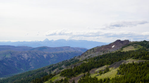 A Mountain View on a partly cloudy day, with a gently sloping mountain with a mixture of grass, rocks, and trees covering the slopes making for an agrarian scene. Just a bit of snow remains near the top of it. 